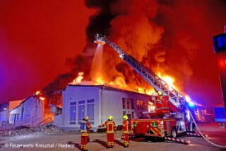 In Kreuztal steht eine Fabrikhalle in Brand. Die Flammen erleuchten hell den Nachthimmel. Die Feuerwehr löscht mit einer Drehleiter von oben.