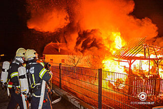 Feuerwehrleute bereiten bei einem Brand in der Ortschaft Freilassing die Brandbekämpfung vor. Es steht eine Halle mit Traktoren in Vollbrand.