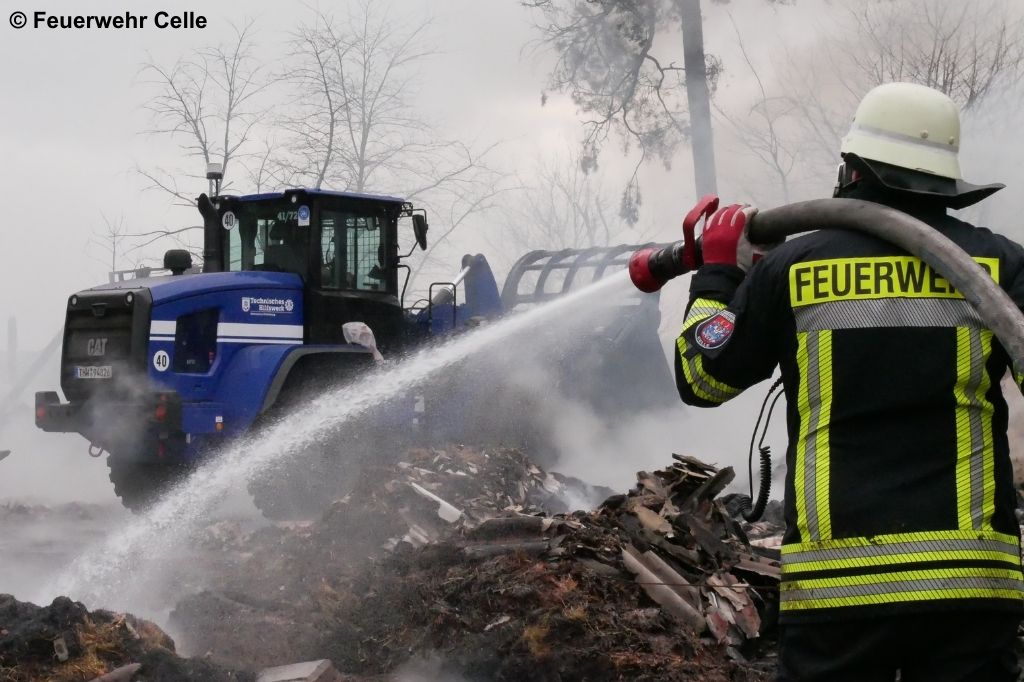 Person in Feuerwehrschutzkleidung mit Helm im Vordergrund richtet einen Wasserstrahl auf einen Trümmerhaufen. Im Hintergrund ein blauer Radlader des THW, der Brandschutt bewegt.